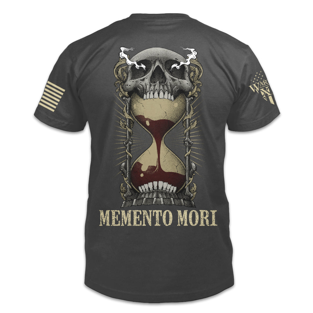 The back of an asphalt-gray t-shirt with an image on the back of an hourglass filled with red sand in the mouth of a human skull and the words "Memento Mori" underneath.