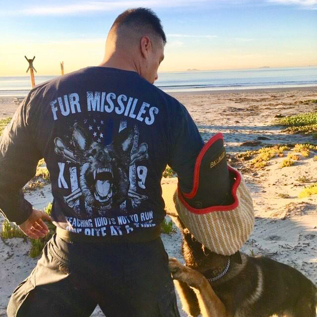 Customer wearing a navy blue long sleeve shirt with the words "Fur Missiles, Teaching Idiots Not To Run, One Bite At A Time!" featuring a german shepherd showing teeth in front of a thin blue line USA flag printed on the back of the shirt.