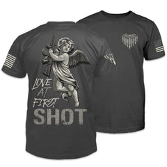 The back and front of a dark grey t-shirt with the words "Love at first shot" printed on the back with an image of cupid holding a rifle, and a small heart made of bullets with the Warrior 12 XII printed on the front pocket area.