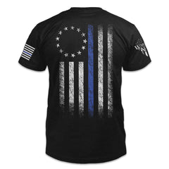 A black t-shirt that features a thin blue line Betsy Ross flag on a back print to show that we remain undeterred in our support for American law enforcement. printed on the back of the shirt.